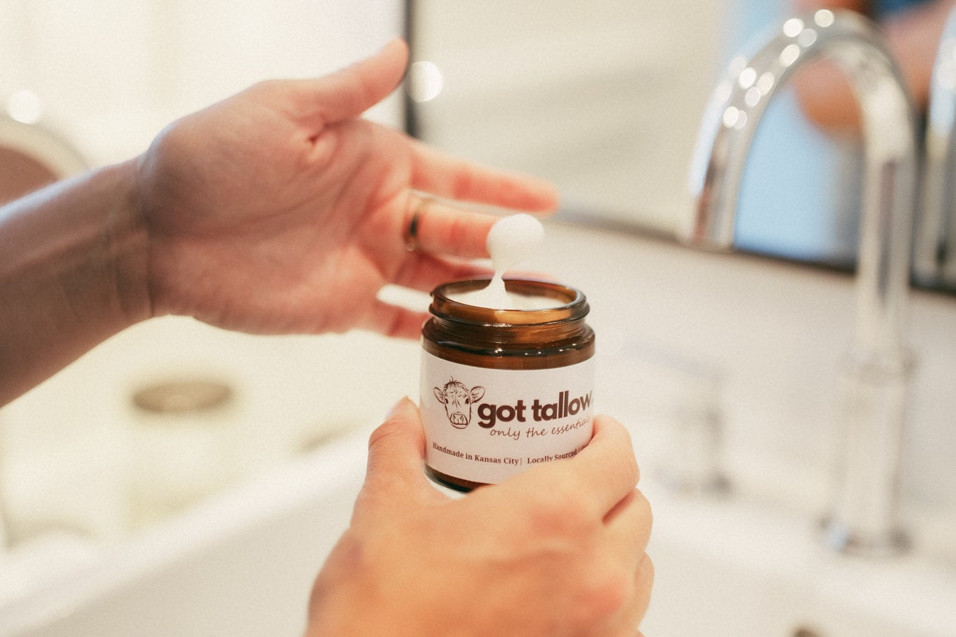 Person applying tallow skincare from a jar labeled 'got tallow' in a bathroom setting.